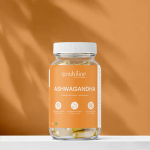 A clear jar labeled Ashwagandha filled with capsules rests on a white surface against a warm orange background. The label highlights benefits such as stress relief and muscle strength.